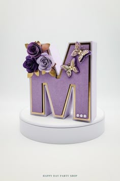 the letter m is made up of purple paper flowers and gold trimmings, along with butterflies