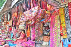 a woman sitting in front of a store filled with lots of colorful fabrics and accessories