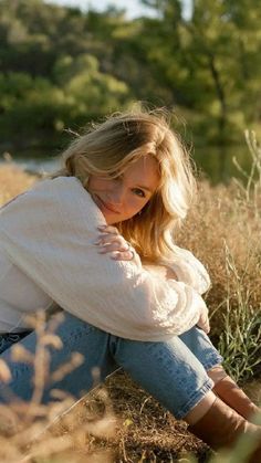 a woman is sitting in the grass with her arms on her knees and looking at the camera