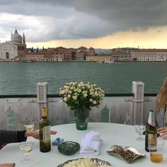 two women sitting at a table with wine and food in front of the water on a cloudy day