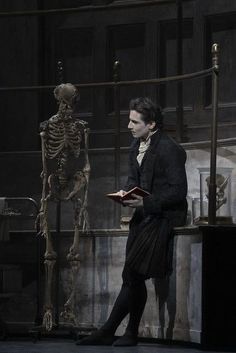 a man standing in front of a skeleton holding a book and looking at the viewer