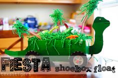 a paper model of a dinosaur in a green wagon filled with plants and trees, on top of a wooden table