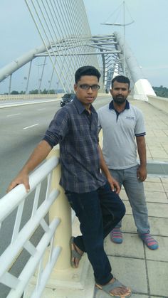 two men standing next to each other on a sidewalk near a bridge with white railings