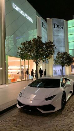 a white sports car is parked in front of a building at night with people walking by