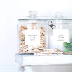 two glass jars filled with clothes pins on top of a white shelf next to a potted plant