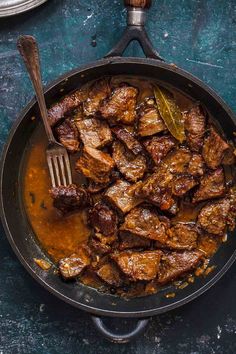 a pan filled with meat and sauce on top of a blue countertop next to two silver spoons