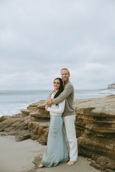 a man and woman hugging on the beach by the ocean with waves in the background