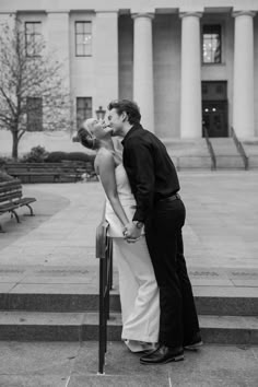 a man and woman kissing in front of a building with columns on the side walk