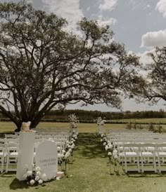 an outdoor ceremony set up with white chairs and flowers on the grass under a large tree