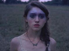 a young woman with blue makeup standing in a grassy field at dusk, staring into the camera