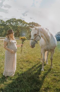 a pregnant woman standing next to a white horse on top of a lush green field