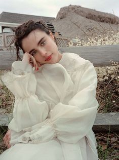 a woman sitting on top of a wooden bench wearing a white dress and holding her hand to her face