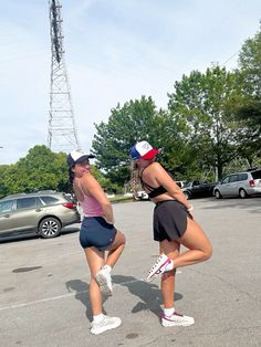 two women in shorts and hats standing in a parking lot with their hands on each other's hips