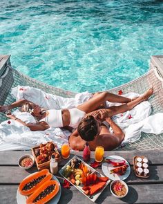 two women laying on a hammock next to the ocean with food and drinks