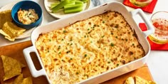 a casserole dish with cheese and vegetables on a table next to chips, beer and wine glasses