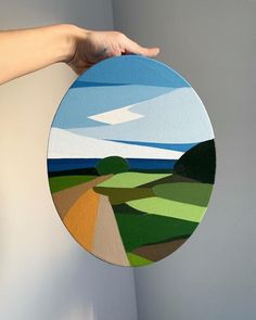 a person holding up a circular painting on the wall with mountains and fields in the background