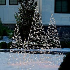 three christmas trees are lit up in the snow with white lights on them and some bushes