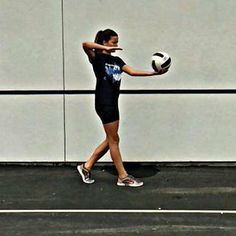 a woman holding a soccer ball on top of a tennis court next to a wall