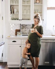 a woman standing next to a little boy in a kitchen with white cabinets and cupboards