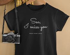 a t - shirt that says sea, i miss you next to a camera on a hanger