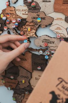 a person's hand pointing at a wooden world map with pins on the countries