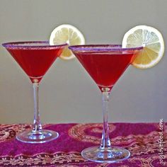 two wine glasses filled with red liquid and lemon wedges on a purple table cloth