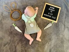 a baby laying on the ground next to some carrots and a sign that says some bunny is 6 12 months old