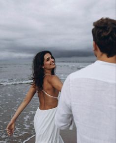 a man and woman are walking on the beach holding hands while looking at each other