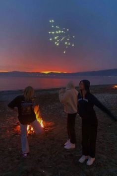 two women standing next to a fire with fireworks in the sky above them at night