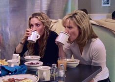 two women sitting at a table drinking from mugs and plates of food on the table