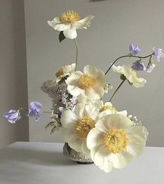 an arrangement of flowers in a vase on a white table top with grey walls behind it