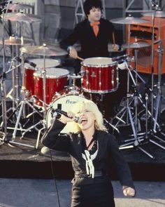 a woman singing into a microphone while standing in front of a band on the street