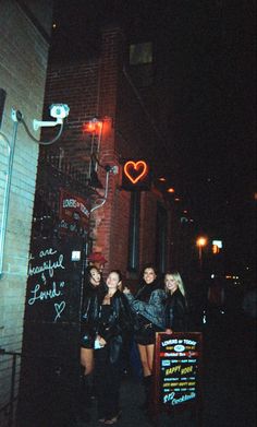 three girls standing in front of a building with a neon heart sign on the wall