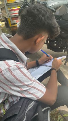 a young man sitting on the ground writing in a notebook with a motorcycle behind him