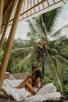 a man and woman sitting on a bed under a palm tree in front of some trees