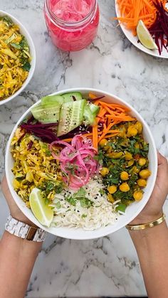 two hands holding a bowl filled with rice and veggies next to another bowl full of vegetables