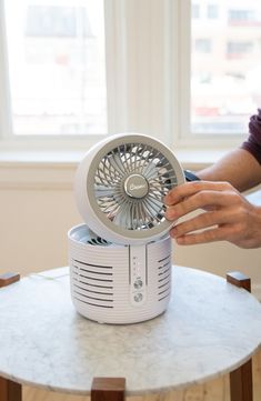 a person is holding an electric fan on top of a small table in front of a window