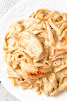 a white plate topped with pasta covered in chicken and sauce on top of a wooden table