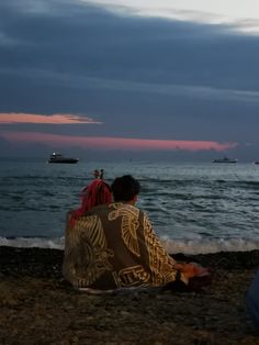 two people are sitting on the beach watching the sun set and boats in the water