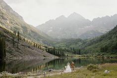 a bride and groom are standing on the edge of a mountain lake