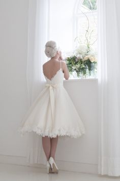 a woman standing in front of a window wearing a white dress with a bow at the waist