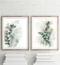 two watercolor paintings hanging on a wall next to white bookshelves in front of a brick wall