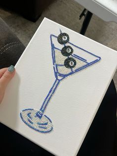 a hand holding up a small canvas with beads on it and a martini glass in the middle