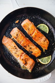 three salmon fillets in a skillet with lemons and seasoning on the side