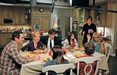 a group of people sitting around a table with food on it in the middle of a kitchen