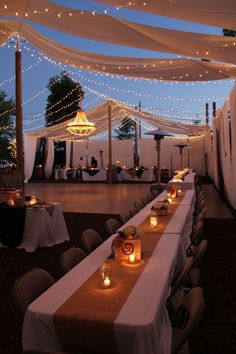 tables are set up with lit candles for an outdoor dinner party at night, surrounded by white draping and string lights