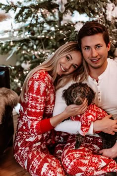 a man and woman sitting in front of a christmas tree with a dog on their lap