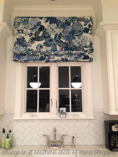 a kitchen window with blue and white floral roman shades over the windows, along with two faucets