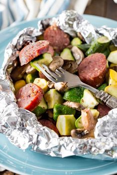 a blue plate topped with foil covered in vegetables and sausages next to a fork