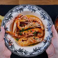 two hands holding a plate of food with shrimp and bread on the table next to it